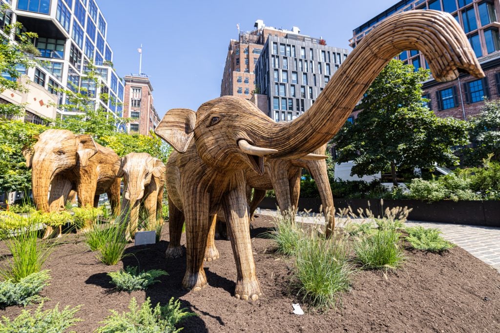 "The Great Elephant Migration" in New York's Meatpacking District. A low-angle photograph of a herd of life-size wooden elephant sculptures amid greenery in front of buildings in the city.