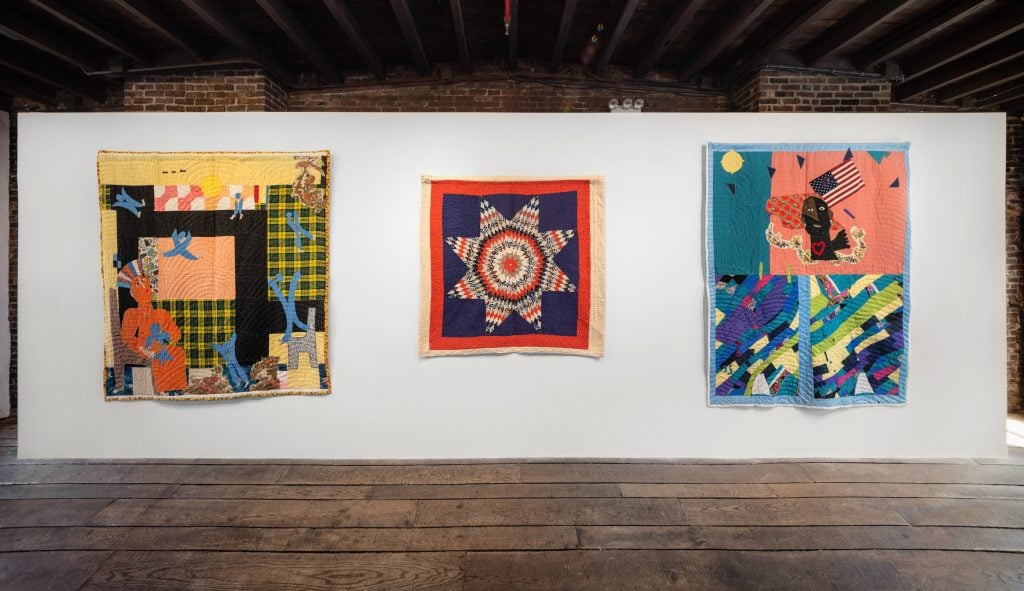 three bright, geometric, and colorful quilts are installed against a white wall in a gallery space with dark wood floors