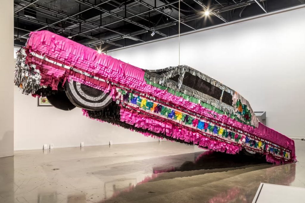 A colorful pink sculpture of a car with tissue paper streaming from it like a pinata