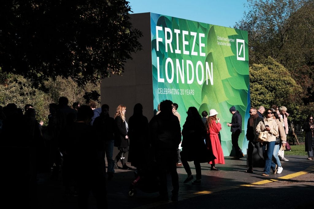 a crowd of people stand outside in a park in front of a large billboard that says frieze london