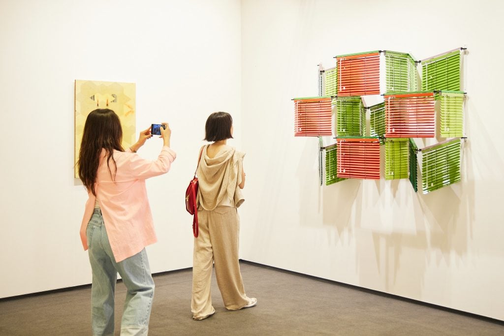 A woman in long dark hair, pink shirt, taking a photo of another woman, short haired, looking at an artwork in green and red on the wall