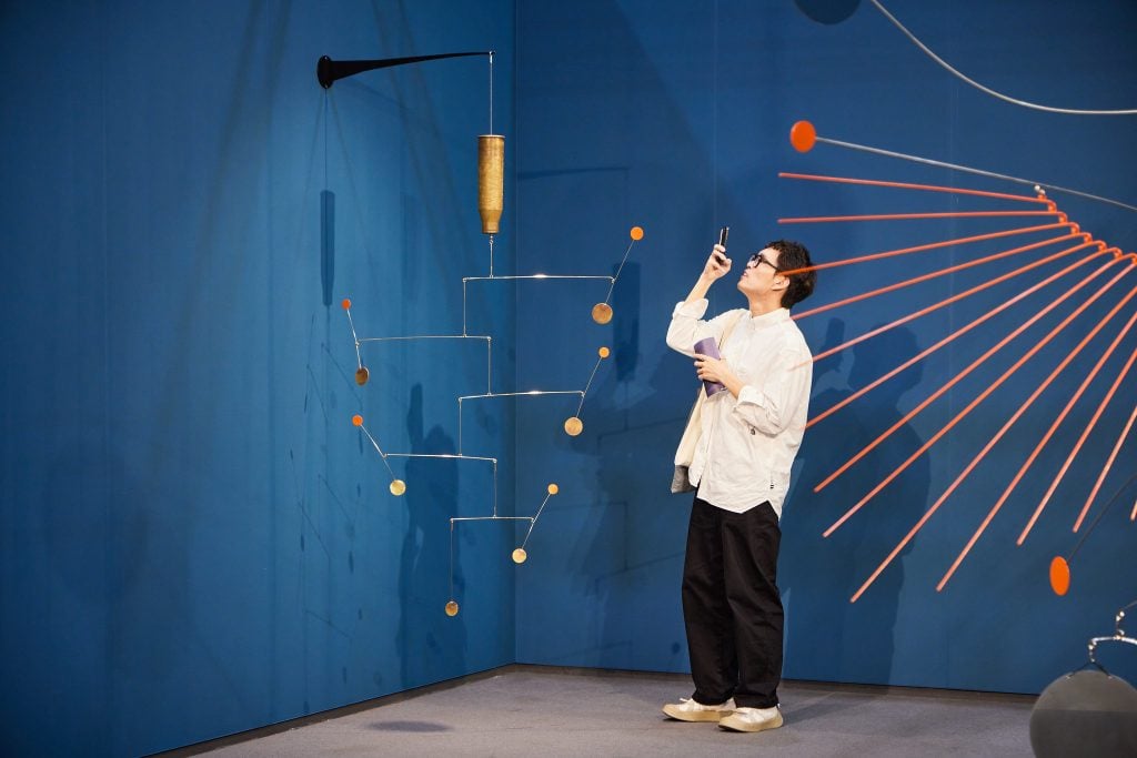 A bespectacled man taking photos of an art work, hung from the ceiling, against a blue background.