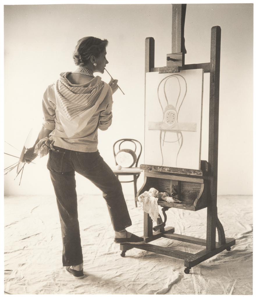 a sepia tone photograph of supermodel Lisa shot from behind while she works stands painting at her easel. The canvas has a chair painted on it