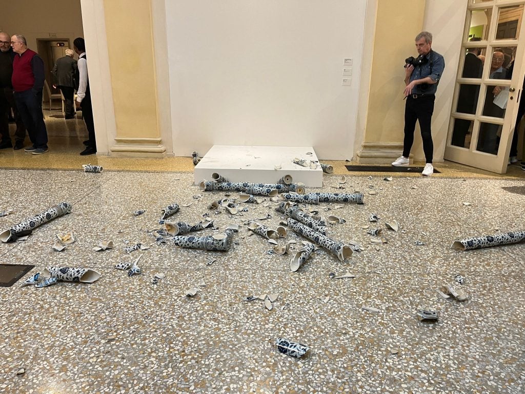 fragments of blue and white ceramics, of which large pieces have a long, hollow tube-like shape, can be seen scattered across a grey stone floor in front of a small raised white podium