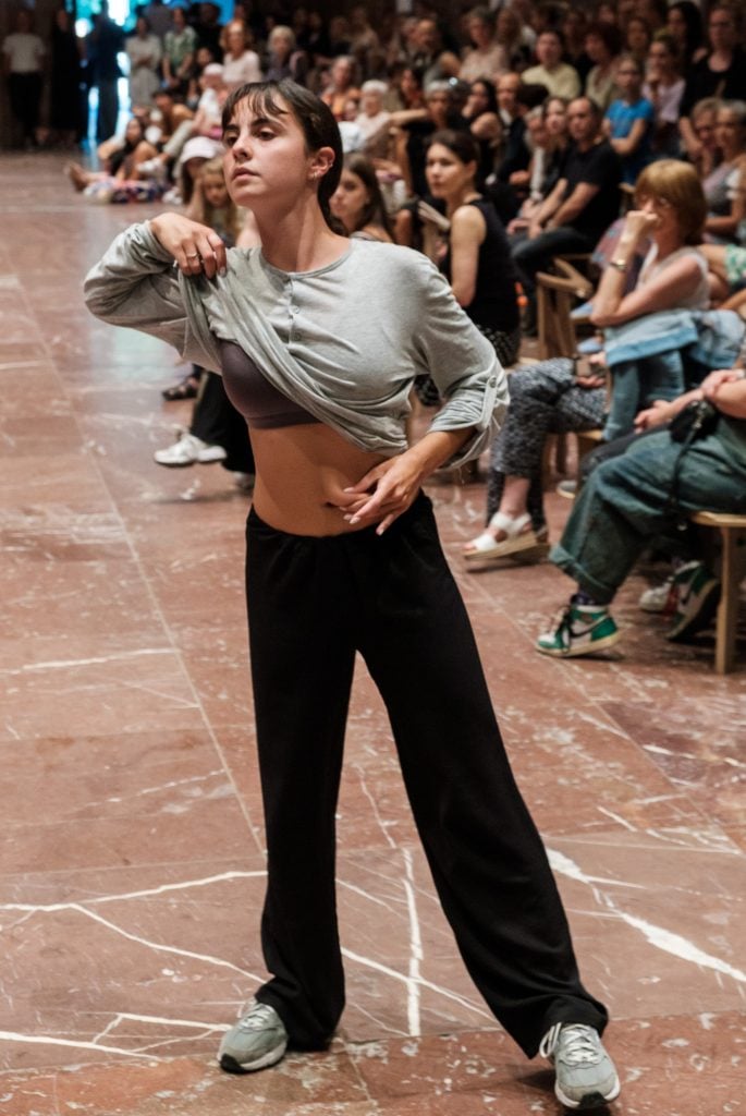 a woman wearing black trouseres and a grey t-shirt appears to be caught in the middle of a swinging motion while she lifts her t-shirt and grabs her stomach and stares into space