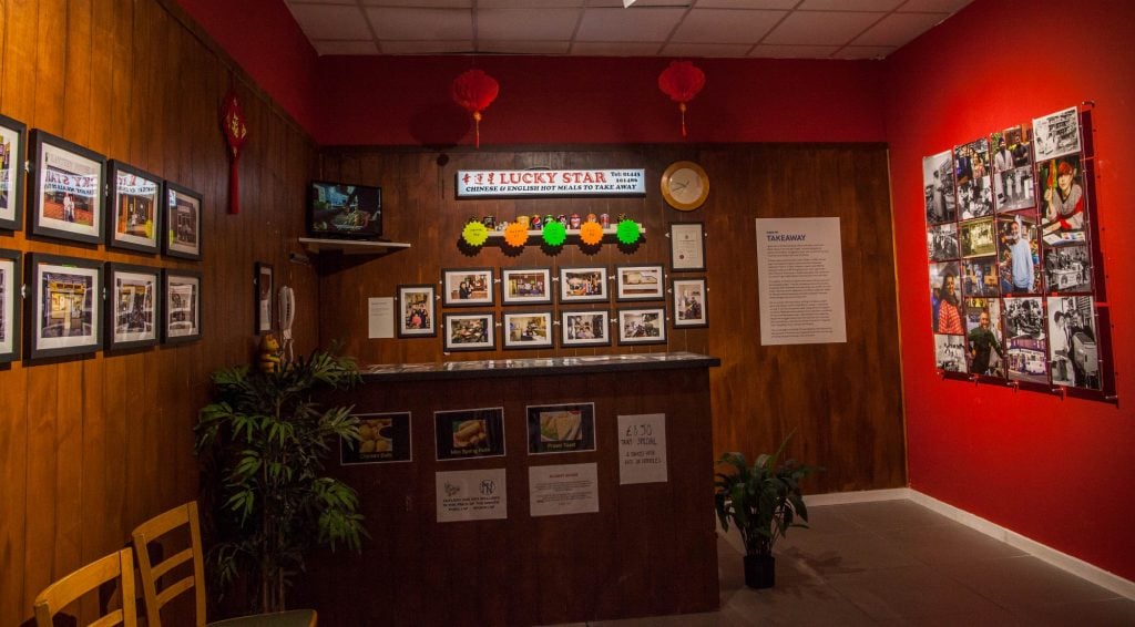 an interior containing a wood counter and wood panelling with menus in a style that is typical of a Chinese takeaway restaurant, a lit-up sign reading "Lucky Star" is affixed to the wall, there are Chinese lanterns and a large house plant