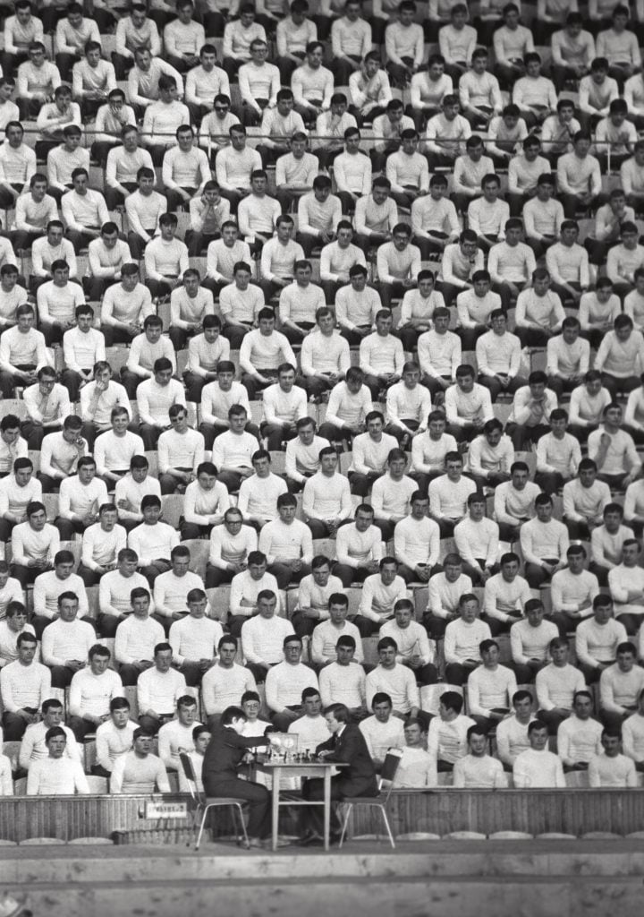 Karpov playing chess in 1978 with a scene of people in white shirts looking on