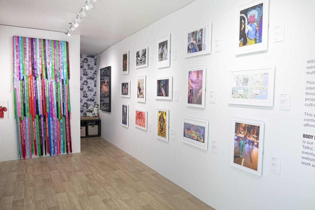 Work on view in the "Body Freedom for Every(Body)" mobile exhibition in Times Square. The interior of a box truck has been transformed into a white wall gallery, with prints hung on one wall, and an Andrea Bowers ribbon installation hanging on the back wall. 