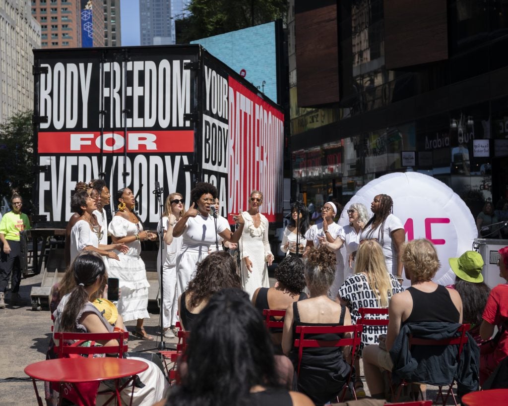 The opening of the "Body Freedom for Every(Body)" mobile exhibition in Times Square. A seated audience watches a performance of female singers dressed in white in front of a large box truck decorated with a black, red, and white Barbara Kruger artwork of the exhibition title. There is also a large inflatable sculpture of an abortion pill. 