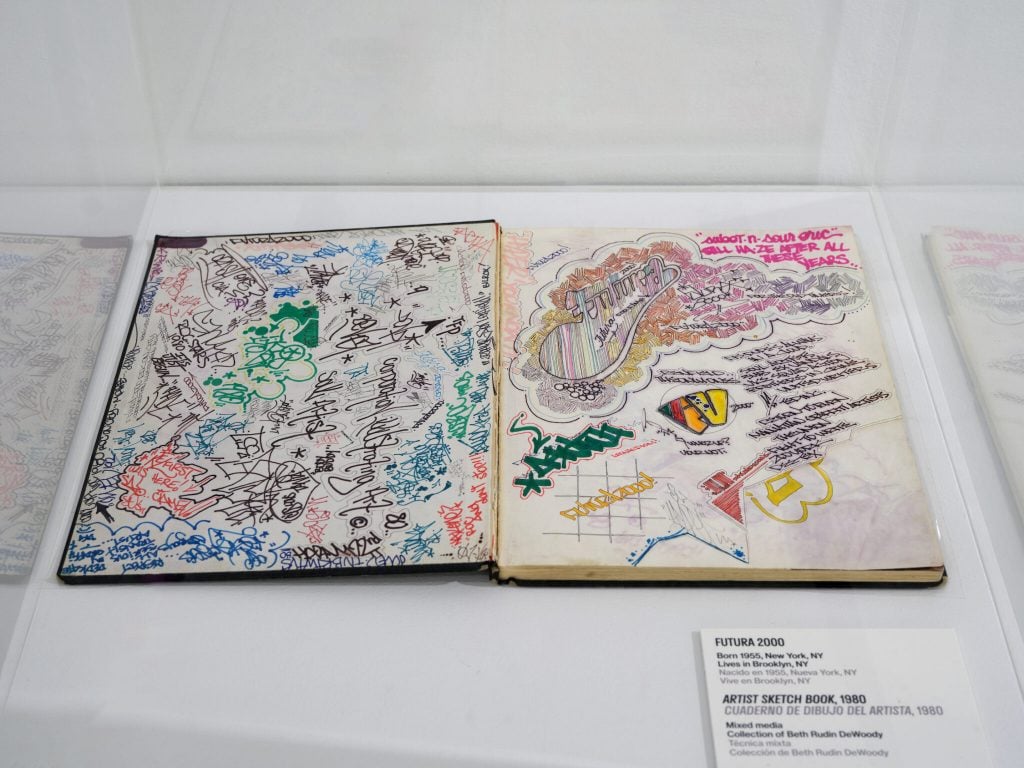 Futura 2000's long-lost black book from 1980, on loan to the Bronx Museum courtesy of collector Beth Rudin DeWoody. A sketchbook in a glass vitrine is flipped open to a page of colorful doodles by graffiti artists.