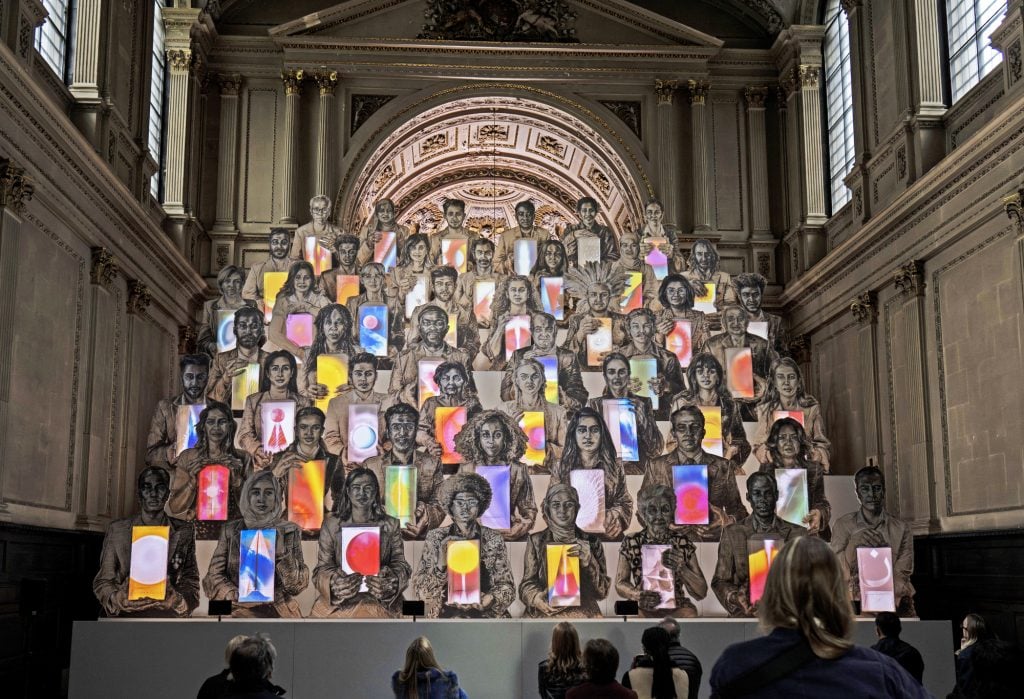 the ornate interior of an old church is filled with a colorful, animated installation in which some 50 drawn figures face us and each hold a box in their hands that contains bright colours like a lava lamp, we can make out the heads of some members of the audience watching this artwork