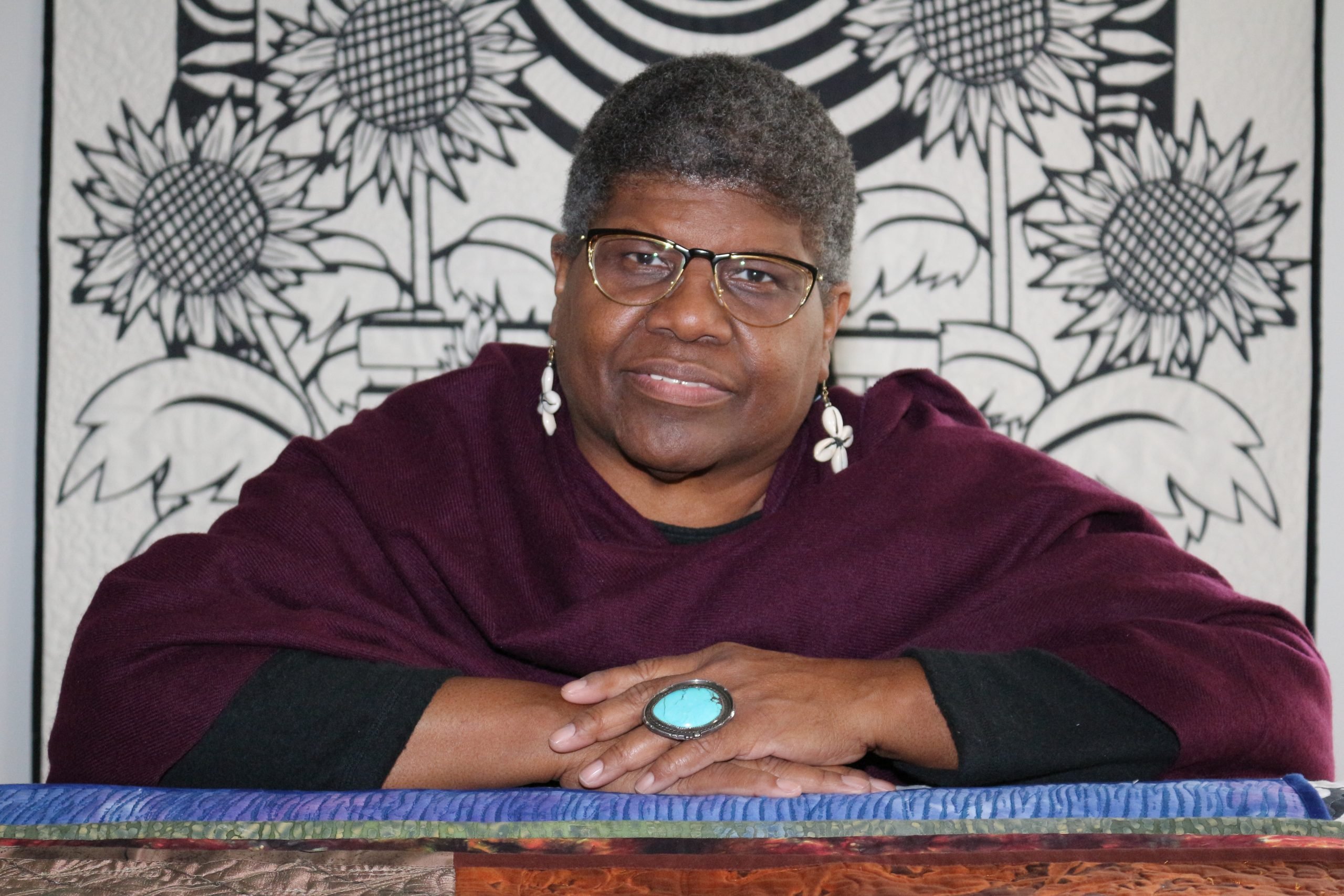 Carolyn Mazloomi, an older place woman with closely cropped graying hair, wearing a long-sleeved purple top, cowrie shell earrings, a large turquoise ring, and gold and black rimmed glasses, sits with her arms on a ledge in front of one of her black and white quilts, which she uses to tell stories of African American history.