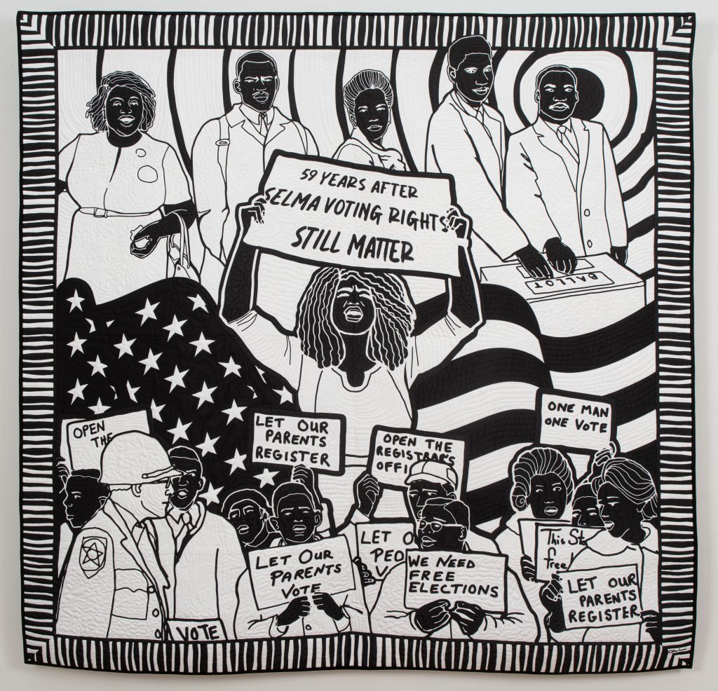 Carolyn Mazloomi Voting Rights. A black and white art quilt loosely divided into three rows. On the top, African Americans line up to cast their vote at the ballot pox. In the center, a Black woman holding a sign reading "59 Years After Selma Voting Rights Still Matter" stands in front of a giant American flag blowing in the wind. The bottom tier shows a crowd of Africans American demonstrating in favor of voting rights, holding a variety of signs, with one white man in a military uniform walking in front of them.