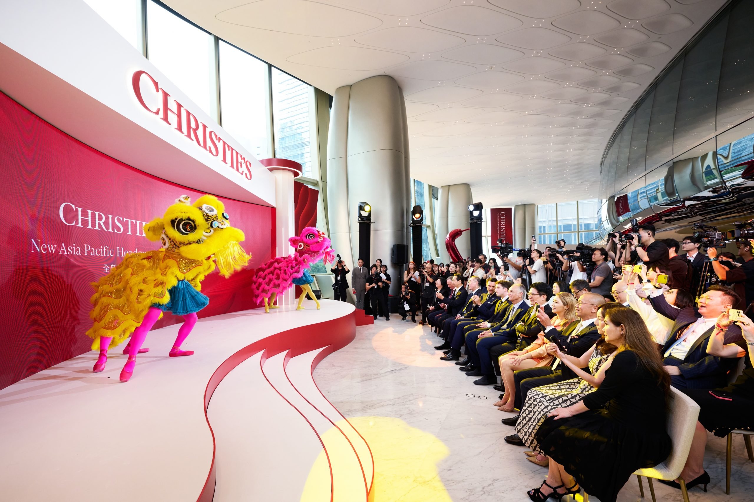 A large crowd applauding Chinese lion dance in yellow and pink on stage, against a red backdrop that spells Christie's