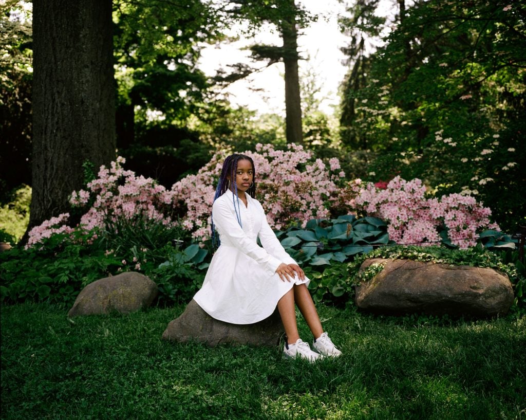 A photo of a black girl in a white dress, sitting on a rock in a park in front of a hedge of pink flowers