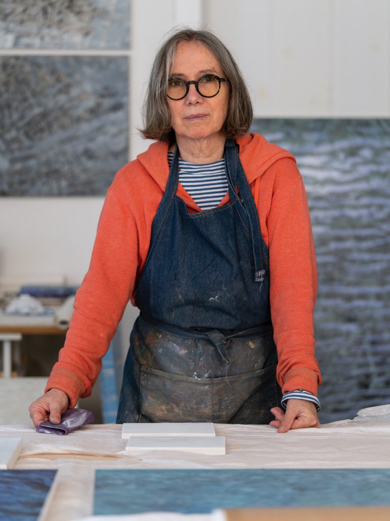 a white woman in her sixties stands at a table in her art studio. she is wearing glasses, an orange hoodie, and a smock.