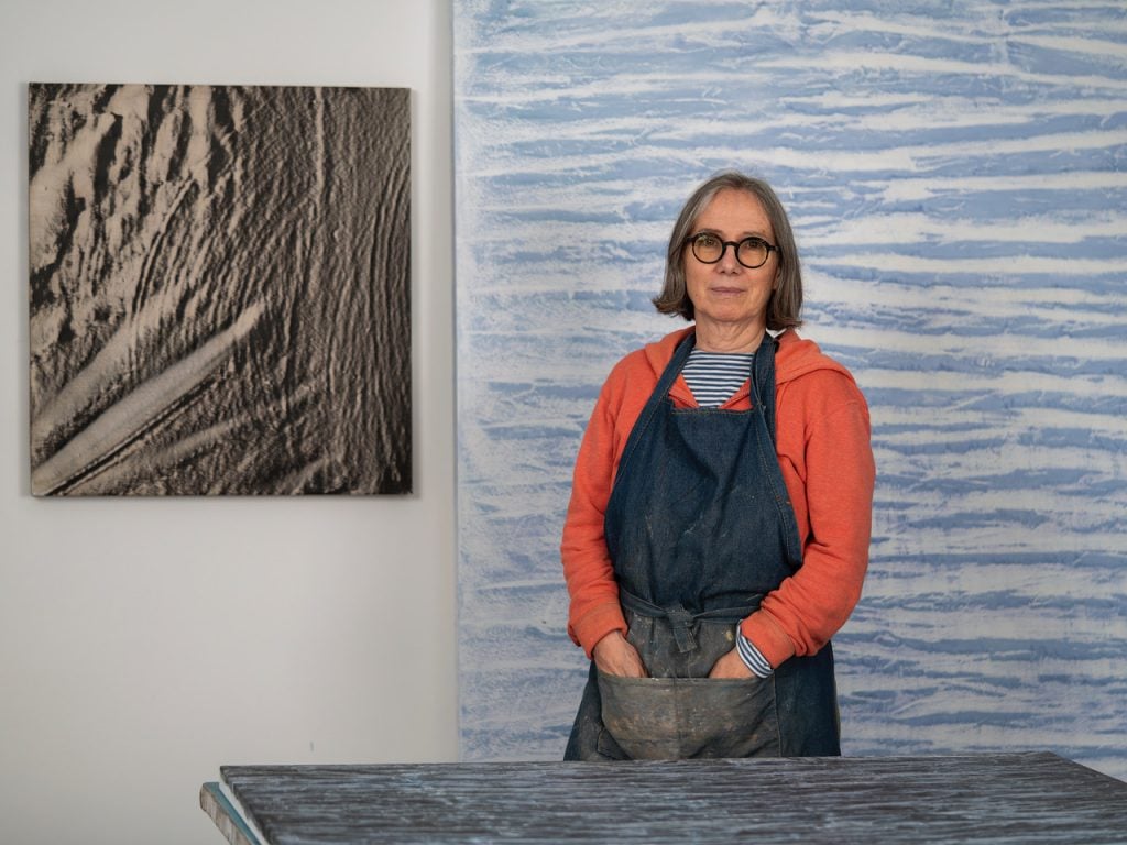 a white woman in her sixties stands at a table in her art studio. she is wearing glasses, an orange hoodie, and a smock.