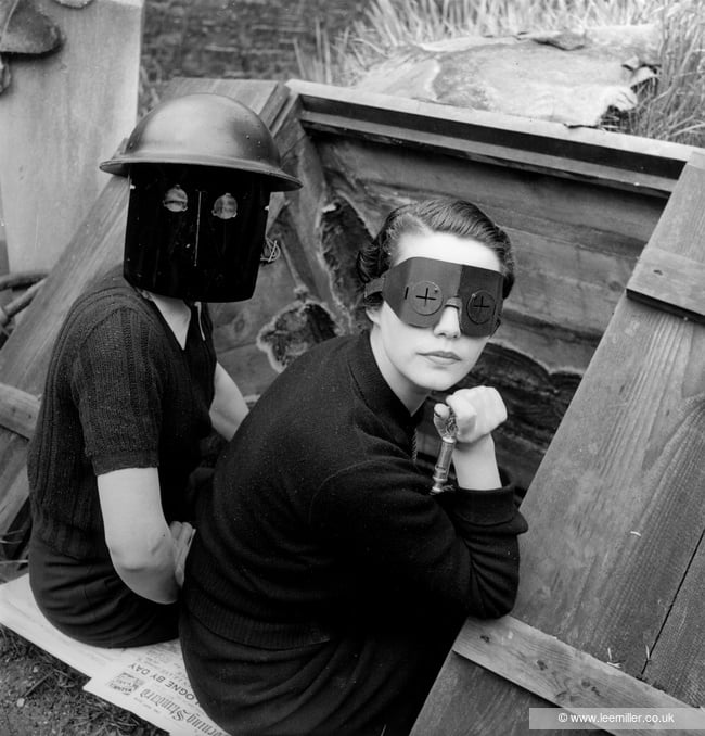 Black and white photo of two women in masks, sitting at the entrance of an underground bunker