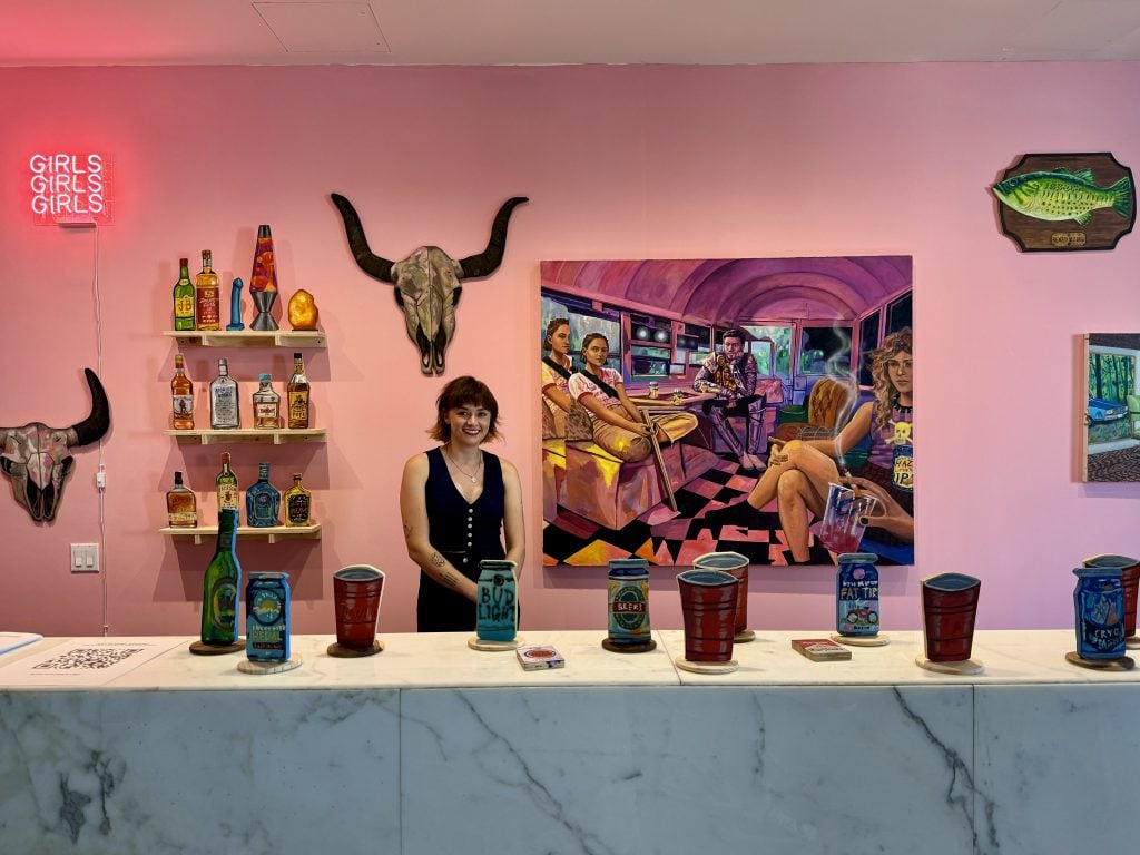 Danielle Klebes with her installation "Just Passing Through," curated by Emily McElwreath, at Spring Break Art Show, New York, 2024. A young woman with chin-length hair stands behind a bar in front of shelves of paintings of liquor bottles, a painting of a bull's skull, and a large painting. 