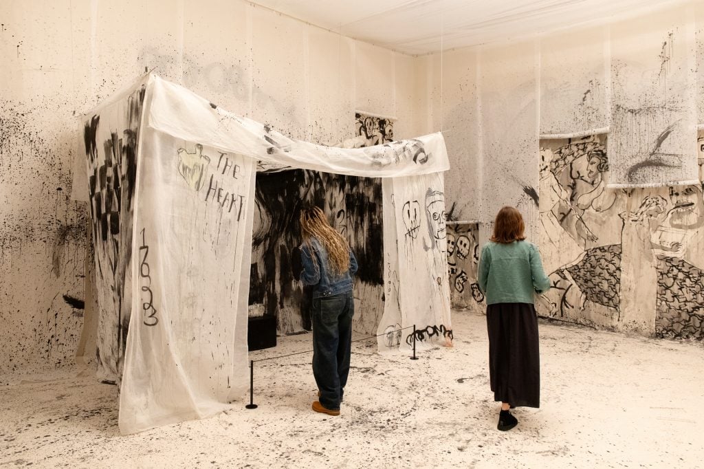 a white fabric tent like structure covered in daubs of black paint, people mill around it and one appears to peer inside to see an soft installation insde