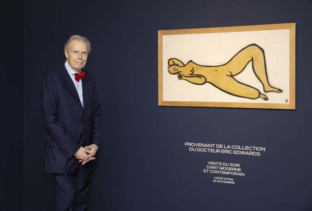 An old white guy in a suit standing next to a nude painting in yellow and milky white colours