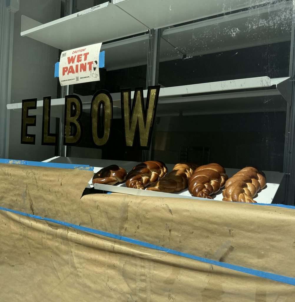 A shope window of a bakery with the words ELBOW in on the glass, challah bread displayed behind it