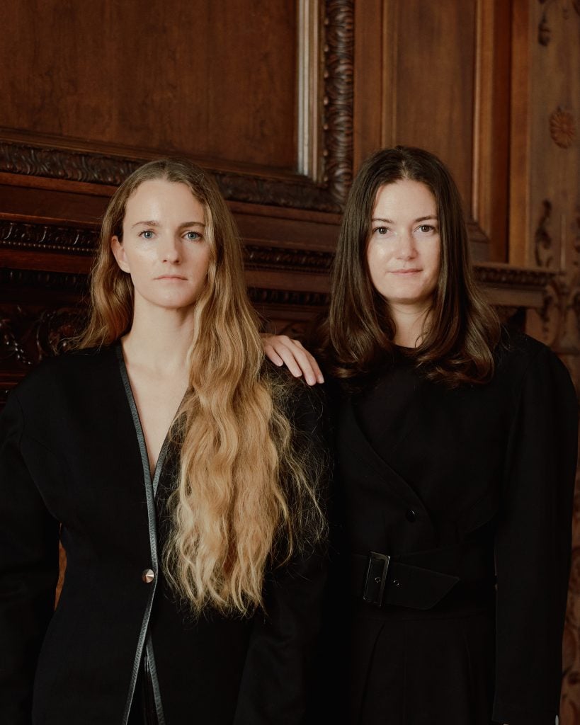 two women Simone Bodmer-Turner and Emma Scully and the gallerist Emma Scully stand against a wooden wall