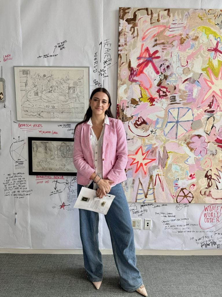 Etta Harshaw, founder of Harsh Collective, with the booth she curated, "All’s Fair in Love and Lore," for Spring Break New York 2024. A young woman in a pink jacket and blue jeans stands in front of a wall with paintings that has been annotated with written notes, like a conspiracy theory white board.