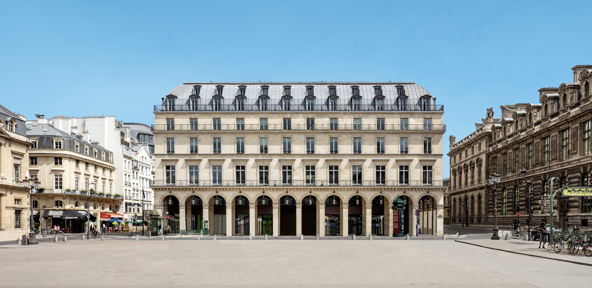 a large classical style building in an urban center, it is positioned on one side of a square and there is blue sky behind