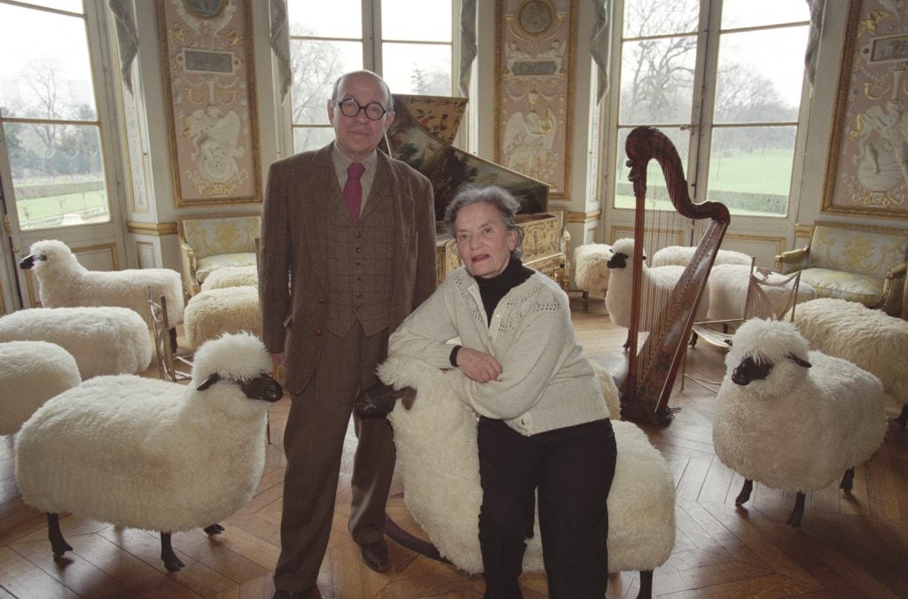 the french sculptor duo Les Lalanne pose sitting on their sheep chairs in a grand room with a harp and piano behind them