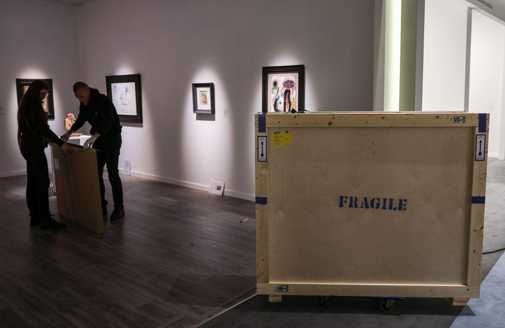 an interior space with artworks hanging on the wall and a large wooden create in the foreground with the word 'fragile' stamped on the front in black typeface, to the left you can see someone opening another similar wooden crate