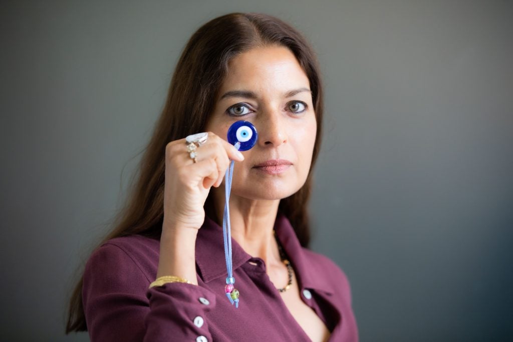 A photo of American author Jhumpa Lahiri in 2022. A middle-aged woman of Indian descent with long dark hair, she is holding up the traditional Nazar symbol of a blue eye 🧿 that is believed to protect against the evil eye in front of her face.