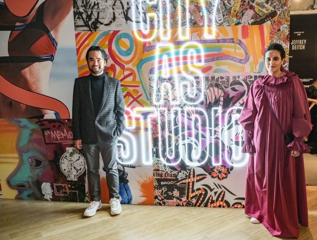 A man in grey clothings and a woman in a purple dress stand in front of a colorful backdrop with flourescent signa