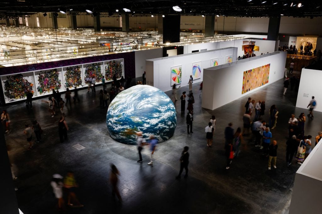 a very large interior space is seen from above partitioned into smaller spaced by white walls that are covered in artworks, installed at the center is a large sculpture of planet Earth