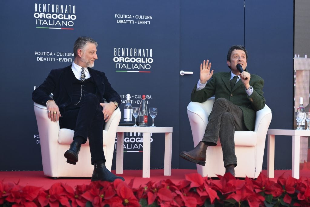 two suited men sit in white armchairs on a stage and one of them is talking into a microphone