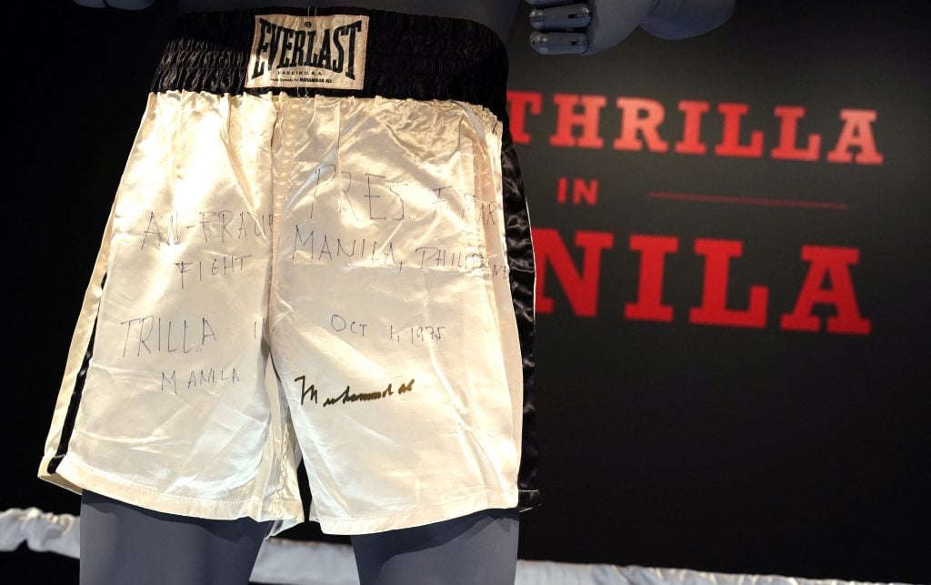 An installation depicting Muhammad Ali's signed satin white Everlast boxing shorts on a mannequin, placed on a mannequin in front of a wall painted with the name of the championship fight Thrilla in Manilla