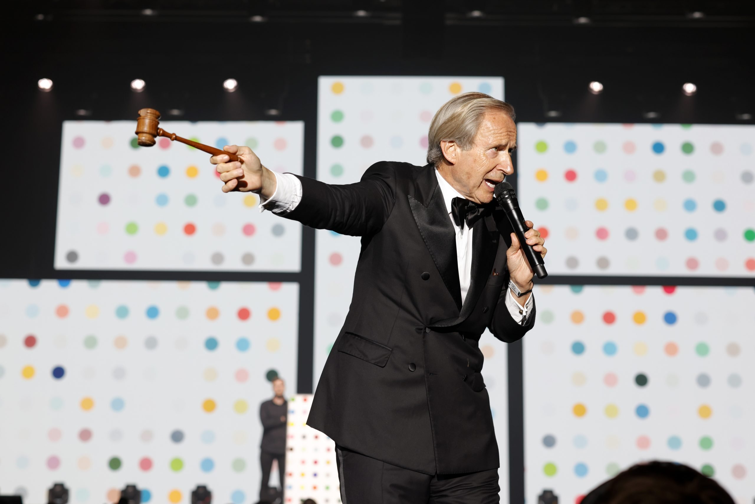 A man in a tuxedo holds a microphone in one hand and a gavel in the other