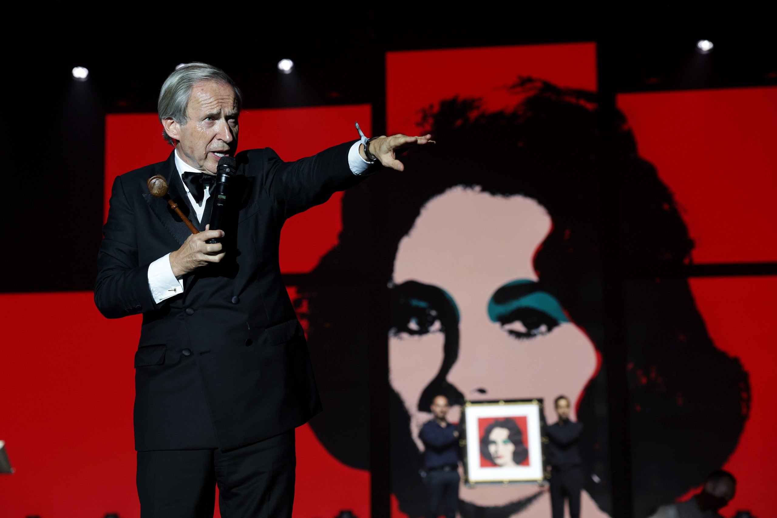 A white man in a tuxedo points from stage in front of a blown-up screenprint pop portrait