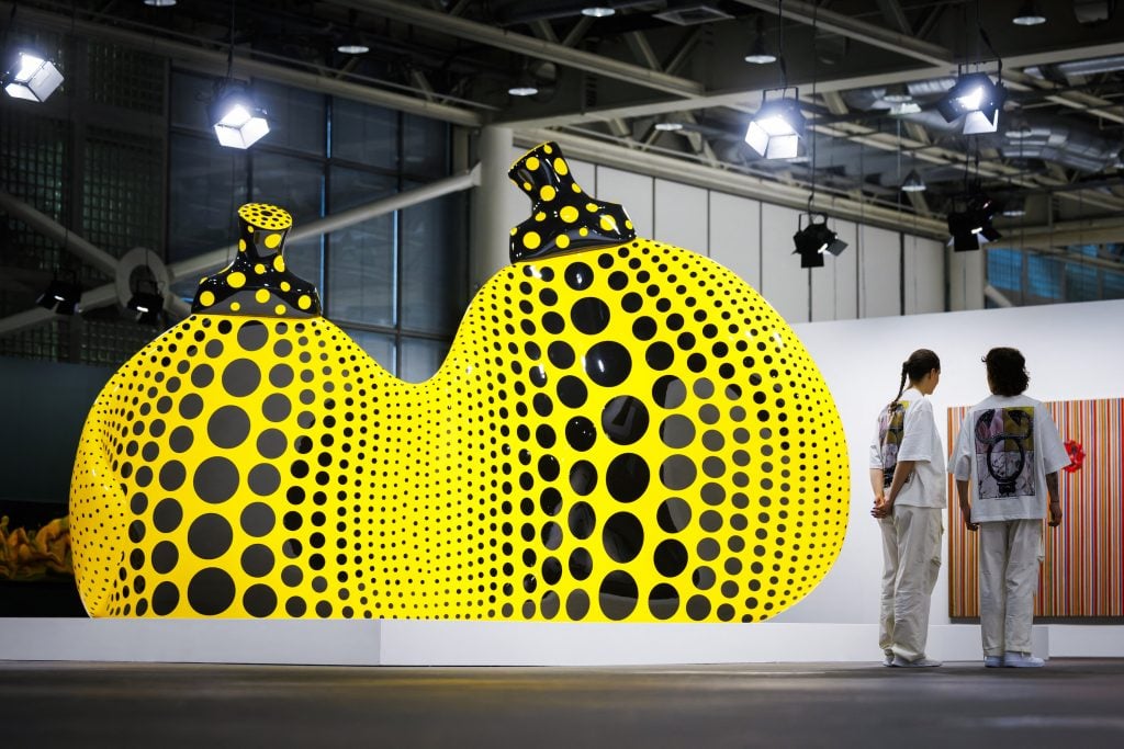 two people stand next to gigantic sculptures of bright yellow pumpkins decorated with black polka dots