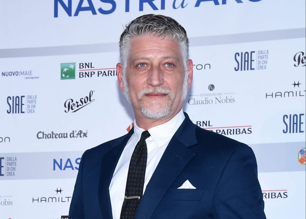 a photograph of a man with grey hair and beard who is wearing a suit and looking into the distance, the photograph is taken at an event and the names of sponsors are seen listed behind him