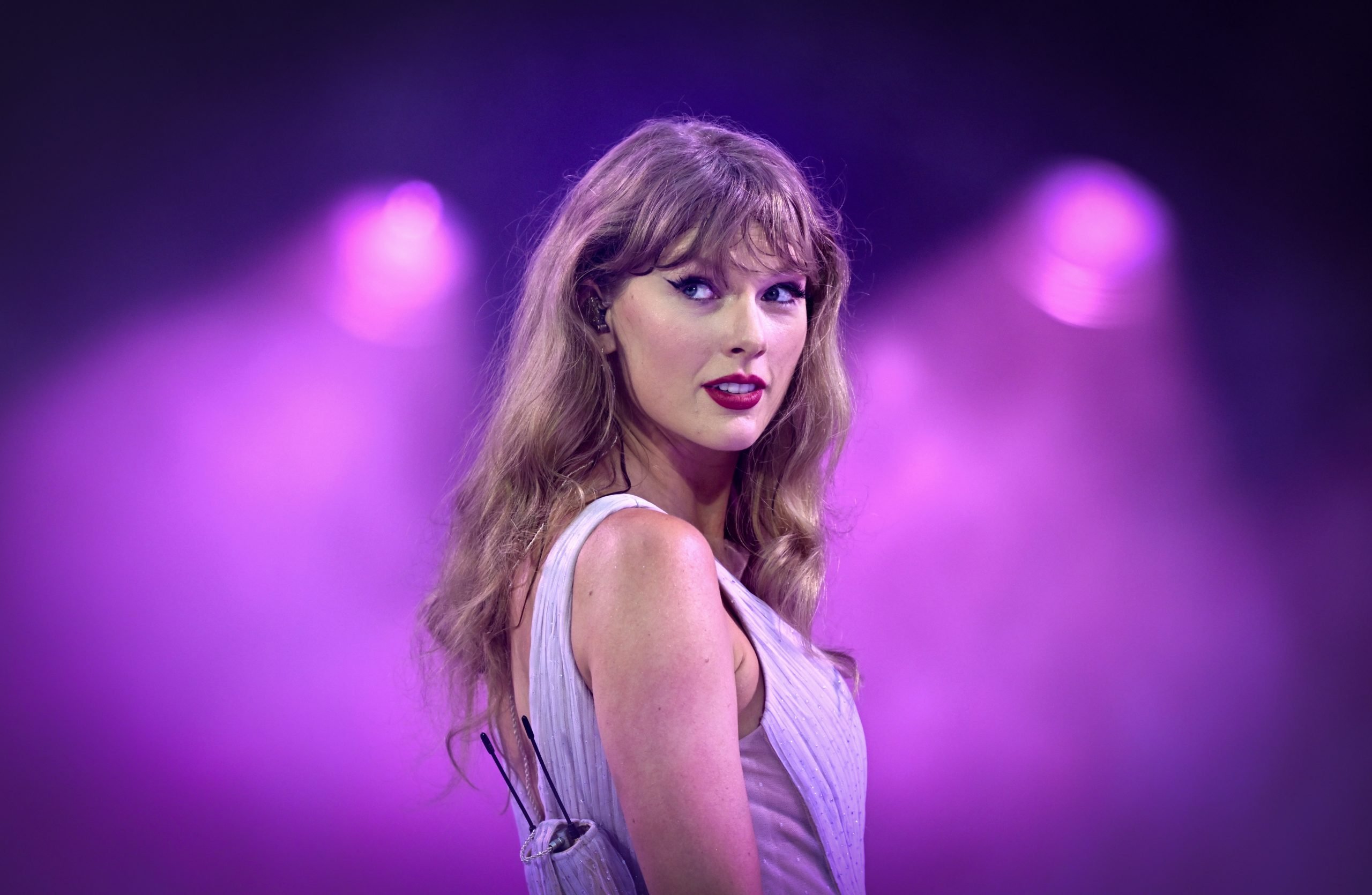 a woman in a purple top against a background of purple lights stares out preciously with long dirty blonde hair, dark lipstick and winged eyeliner