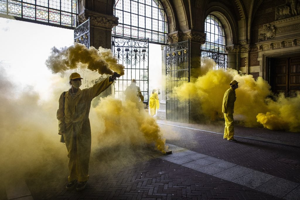 two people wearing all yellow jumpsuits hold up canisters releasing dense yellow smoke into the air, they are standing inside an old-fashioned, spacious interior space with gates and large glass windows