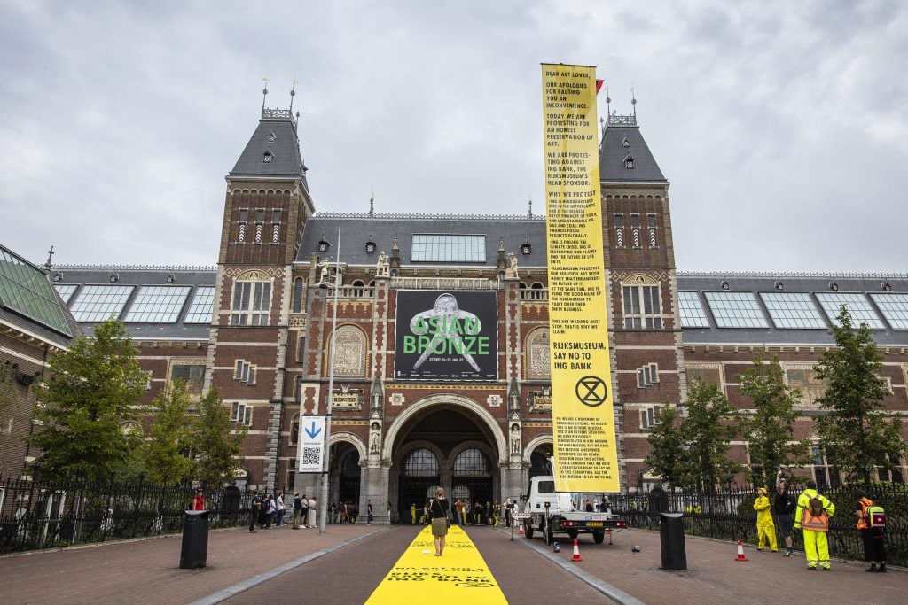 A tall yellow banner rises to the right of the facade of a large stately building against a grey sky, the banner has chunks of text on it in black