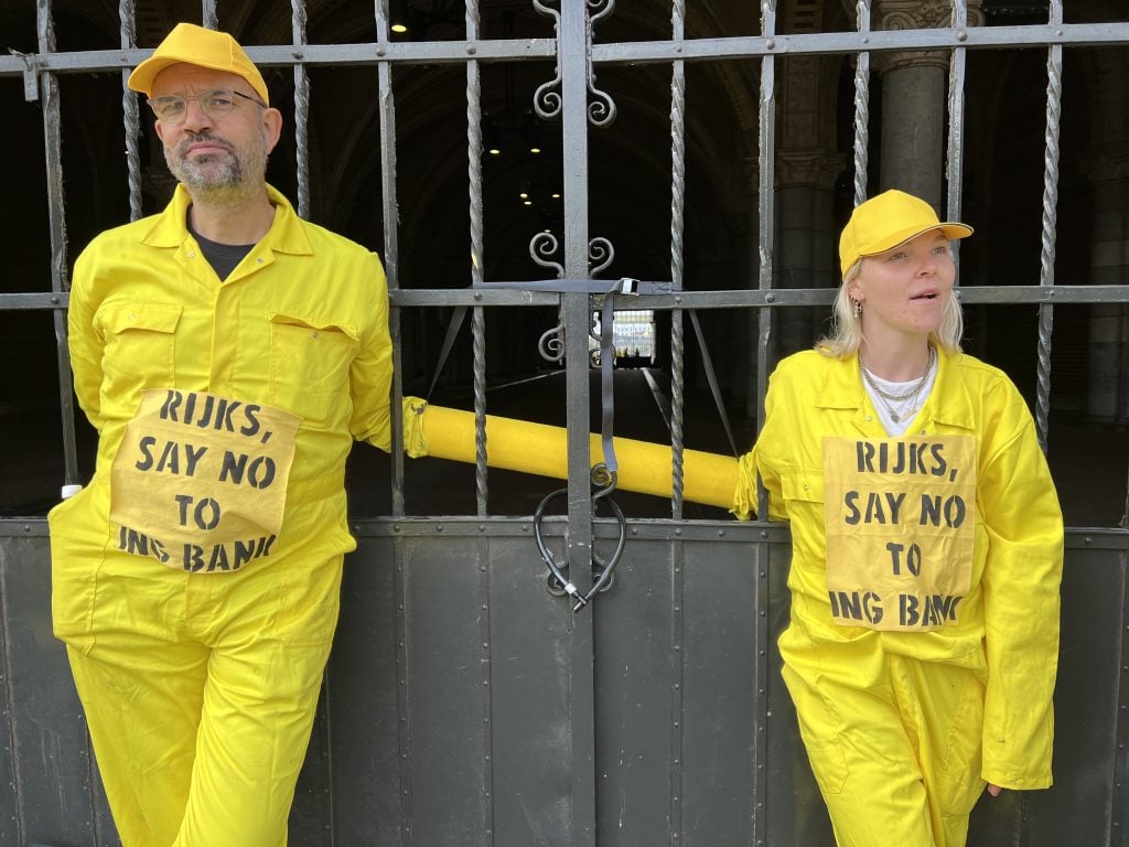 a man in a woman wearing yellow jumpsuits and caps with "Rijks Say No To ING Bank" pasted onto their chests stand chained to the railings on a large entrance gate