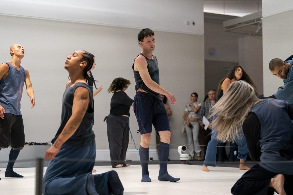 a group of people in blue sweatpants and shirts move around in a gallery space as people look on