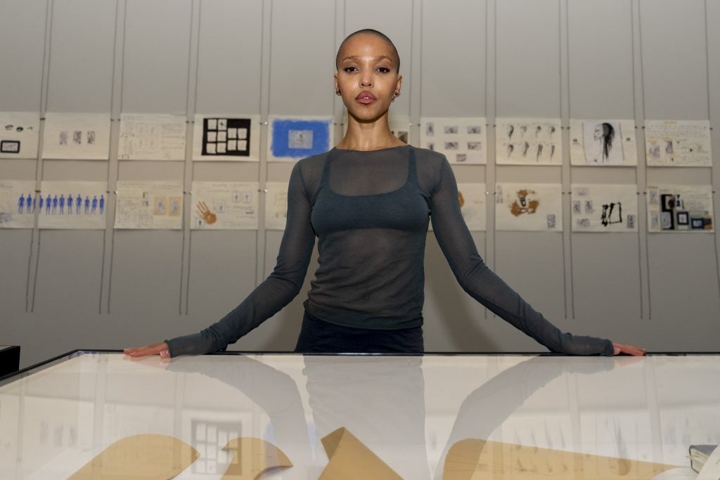 a woman stands in front of a series of small drawings on paper on the wall behind her