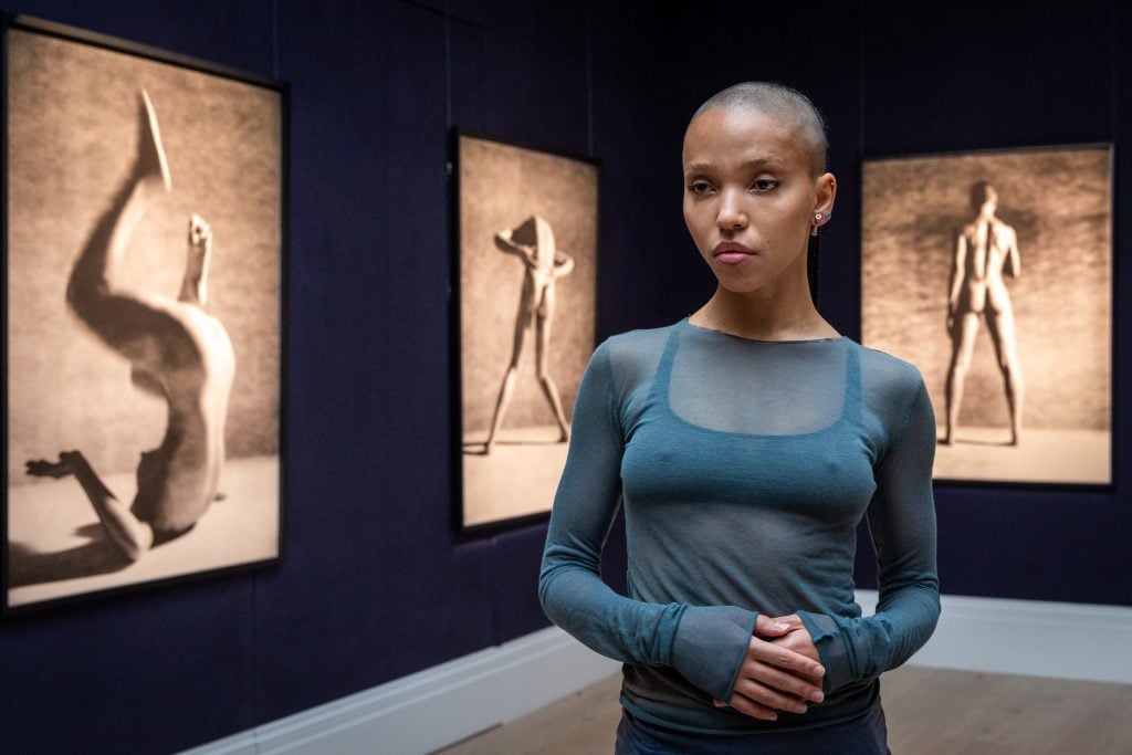 A young black woman in a blue top stands amid brown-tinted portraits of a nude figure