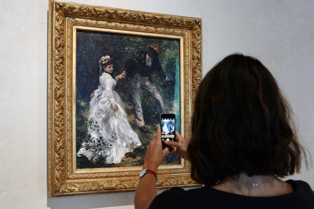 The back of a woman's head is visible as she takes a cell phone photo of an impressionist painting of a woman in a white dress and a man