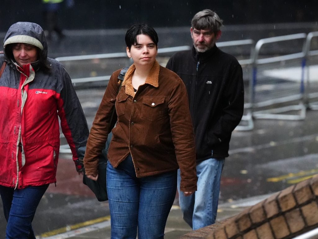 a group of people walk along the street, of which the foremost figure is a young woman with dark hair in a bob and a brown jacket and jeans