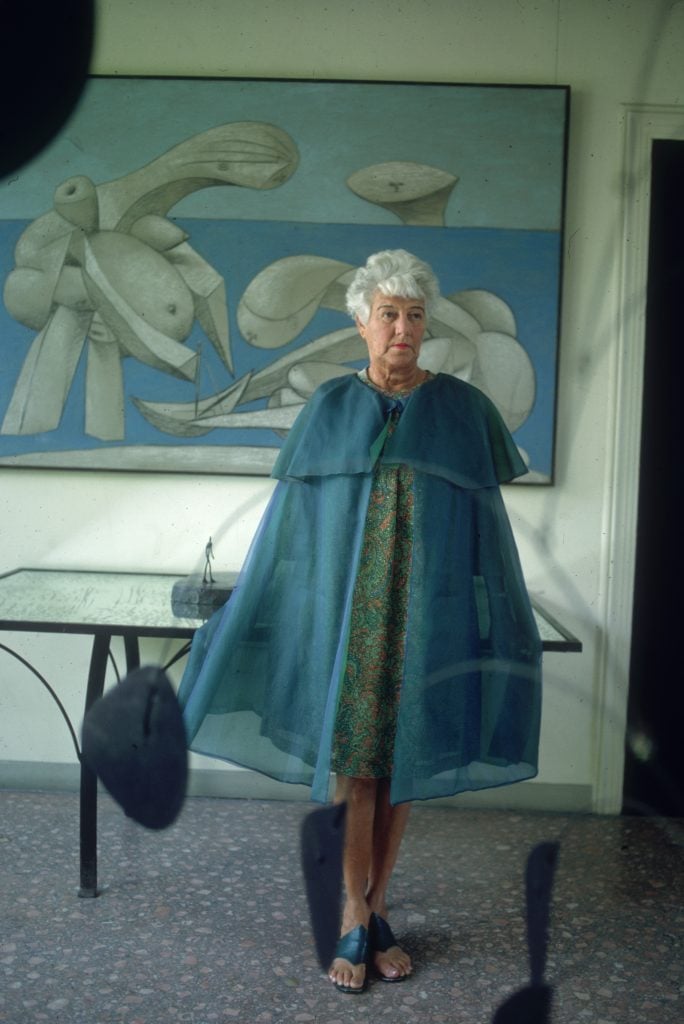 A woman with white hair wears a dramatic blue cape over a dress in front of a painting